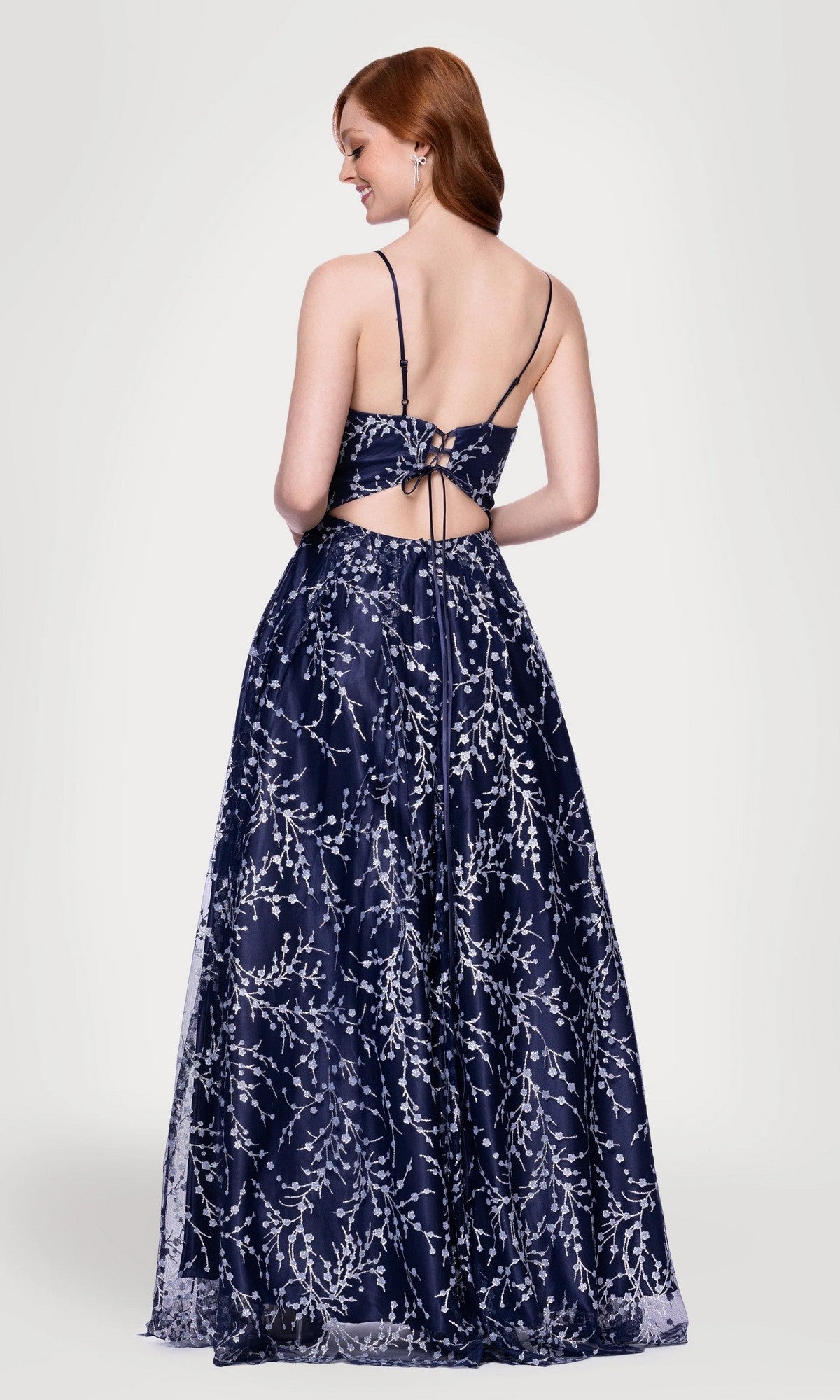 A woman models the Blondie Nites 5386BN long prom dress—navy blue, floor-length with an A-line skirt, silver floral glitter print, open back, and thin straps—against a plain light background.